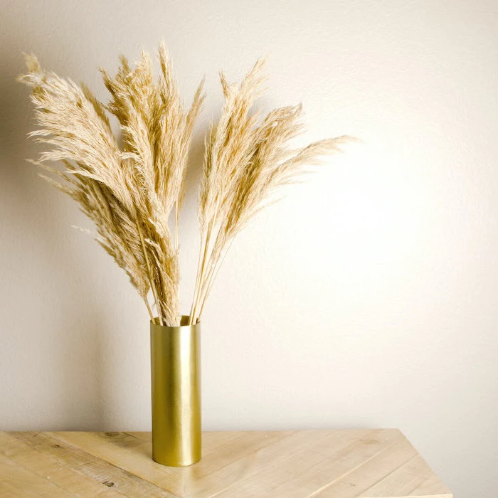 gold vase with tall pampas grass inside placed on wooden table photographed on white background