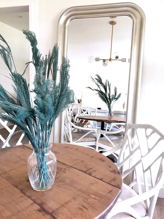 green tall pampas grass inside glass vase placed on wooden table with white chairs large mirror next to it leaning on white wall