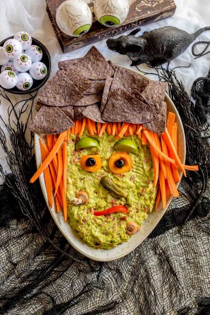 guacamole dip arranged as witch face halloween party food for adults baby carrots black tortilla chips around it