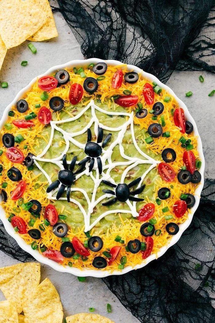 halloween appetizers guacamole dip on top of taco bake grated cheese chopped olives cherry tomatoes for garnish spider web spiders made of olives