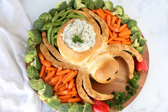 halloween party appetizers bread baked in the shape of spider baby carrots peas broccoli arranged around it on wooden cutting board