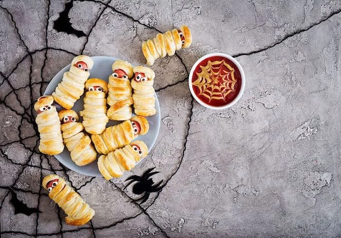 halloween themed food sausages wrapped in dough as mummies arranged on grey plate tomato dip in white bowl placed on gray surface