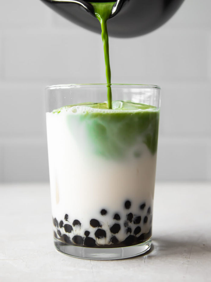 matcha tea being poured into glass filled with milk and tapioca pearls how to make boba tea placed on white surface