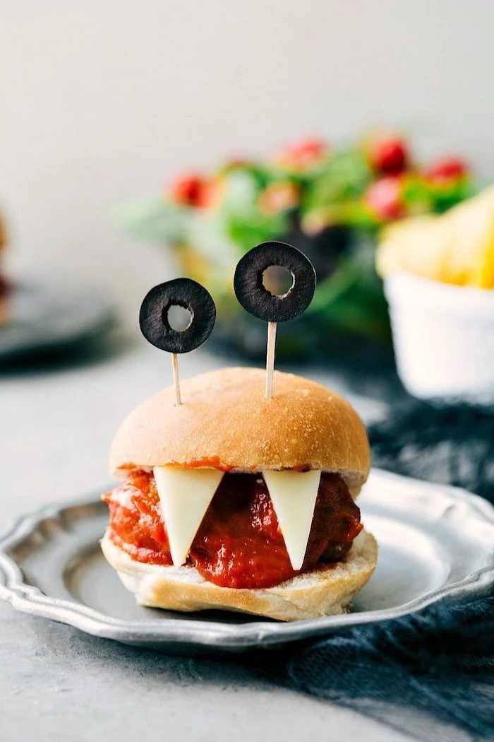 monster burger with teeth made of cheese olives for eyes halloween finger foods placed on gray metal plate