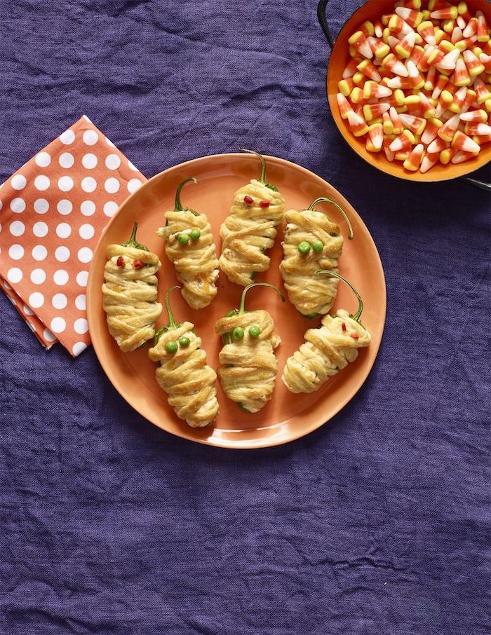 mummy peppers wrapped in dough arranged on orange plate halloween appetizers candy corn in orange plate purple table cloth