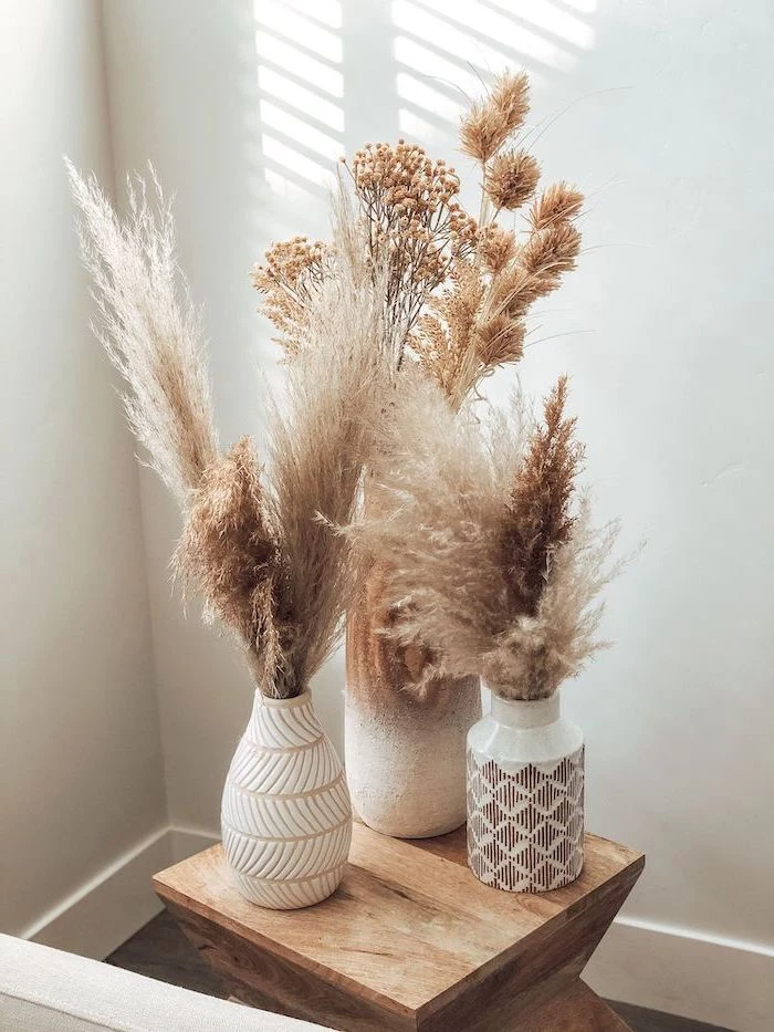 three ceramic vases filled with pampas grass placed on small wooden table white wall in the background