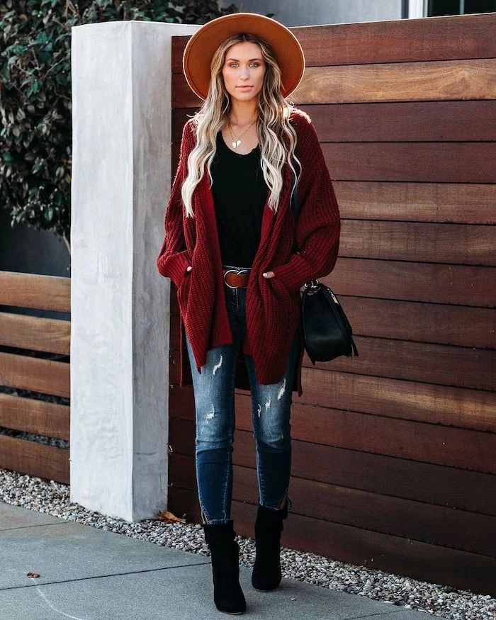 woman with long blonde wavy hair womens fall fashion wearing jeans black top red cardigan black boots and bag standing on the sidewalk