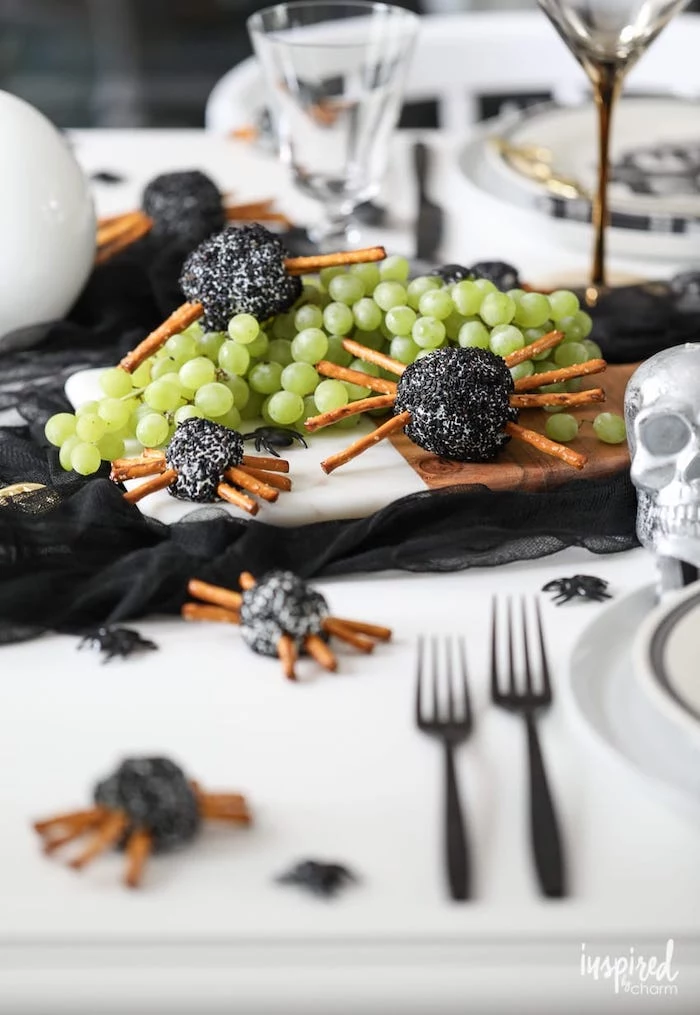 wooden cutting board placed on table with black tulle on it halloween party appetizers cheeseballs with pretzels arranged as spiders