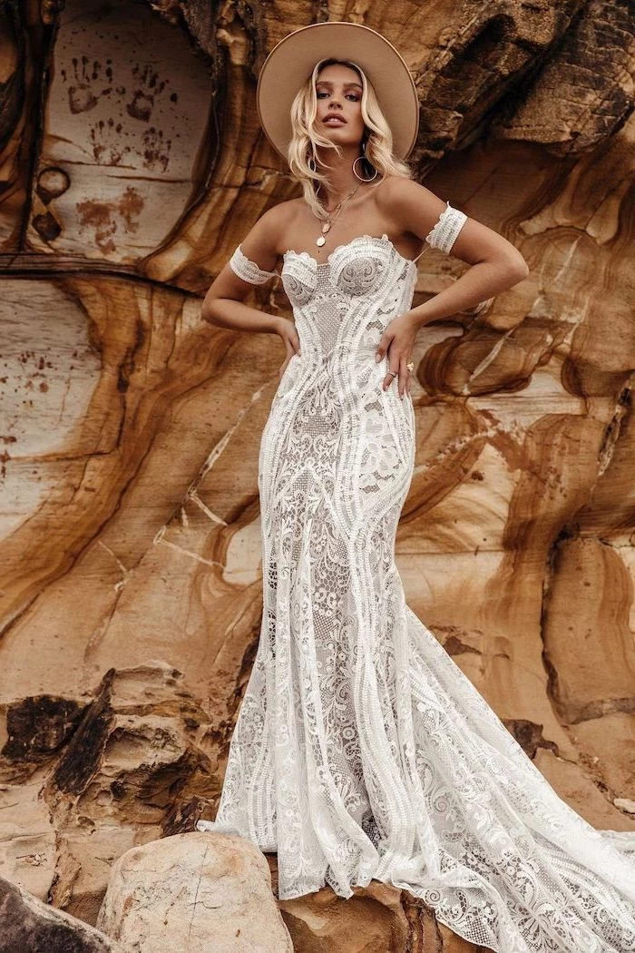 blonde woman wearing all lace dress with heart shaped neckline bohemian wedding dress wearing a baige hat standing on rocks