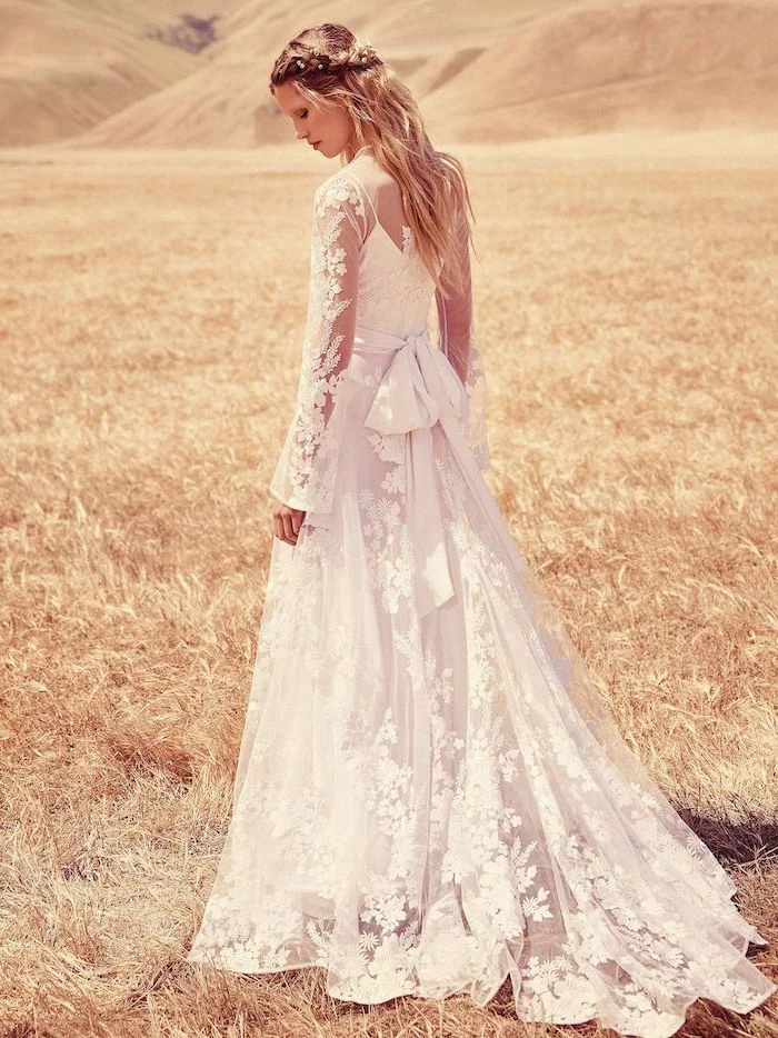 blonde woman with long wavy hair wearing lace boho wedding dress with long sleeves standing in the middle of field