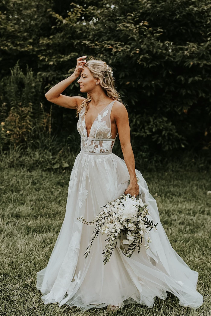 blonde woman with wavy hair wearing dress made of tulle with lacy flowers on it boho wedding dress holding white flower bouquet