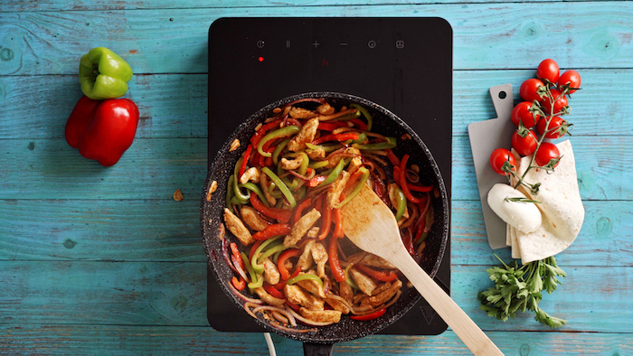 chicken cooking with onion and peppers in black saucepan with wooden spatula on blue wooden surface traditional mexican food