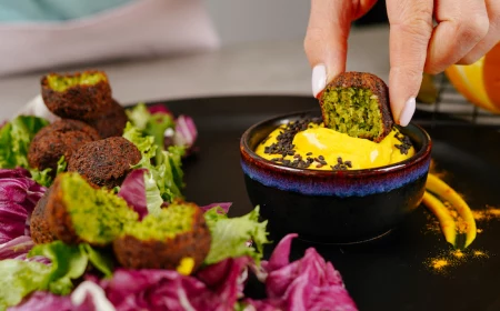 half of falafel dipped into yellow dip with black sesame seeds vegan finger food black dip bowl placed on black plate