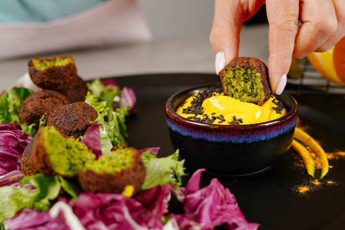 half of falafel dipped into yellow dip with black sesame seeds vegan finger food black dip bowl placed on black plate