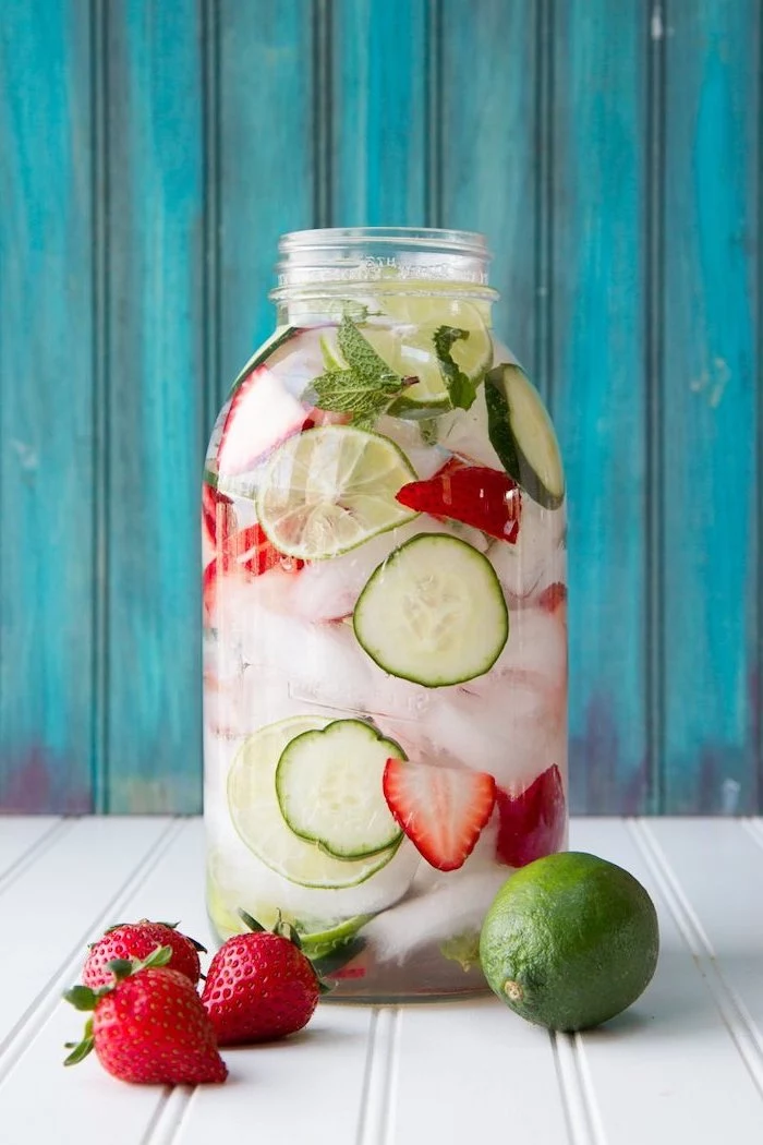 large jar filled with cucumbers lime strawberries mint leaves best detox drink placed on white wooden surface