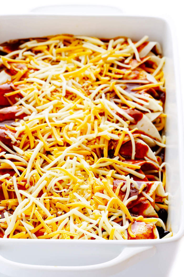 mexican food recipes enchiladas covered with tomato sauce different kinds of shredded cheese placed in white baking casserole dish