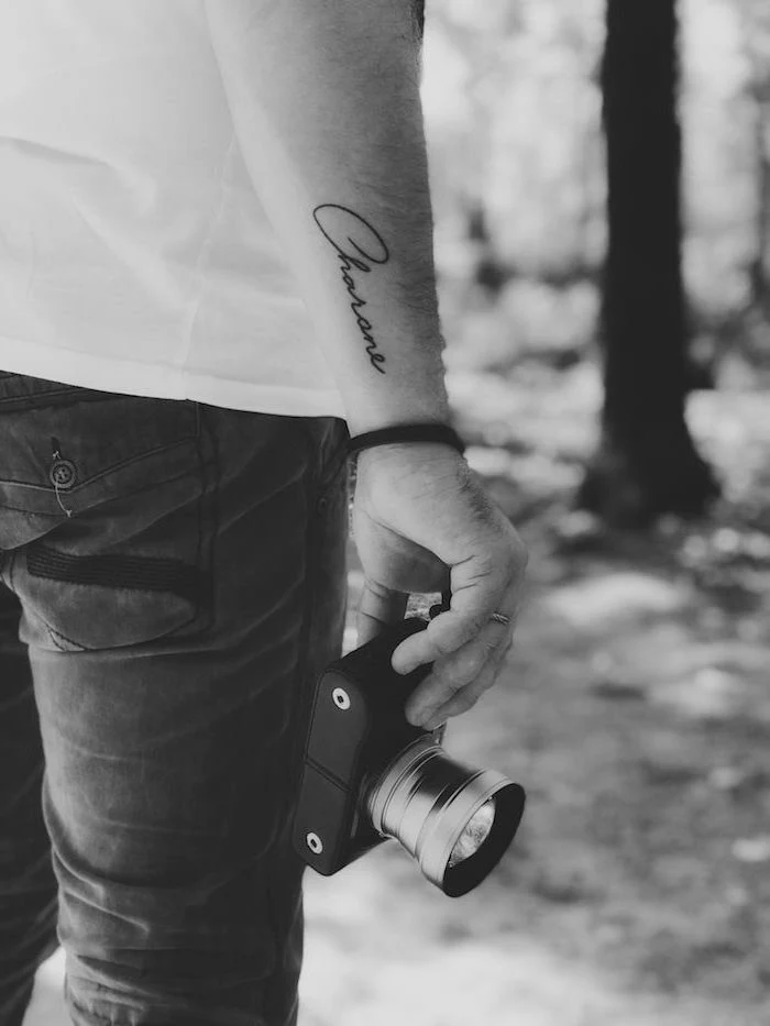 name written in cursive font on the side of the wrist on man holding camera small tattoo ideas for men black and white photo