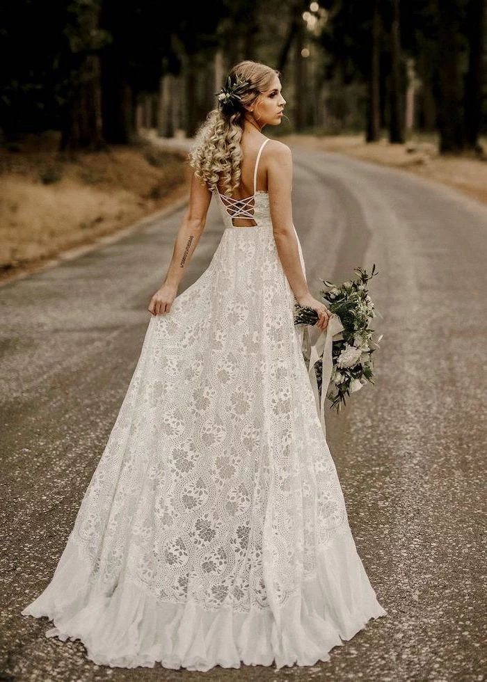 photo taken on lone road with trees unique wedding dresses blonde woman wearing all lace white wedding dress holding white flower bouquet