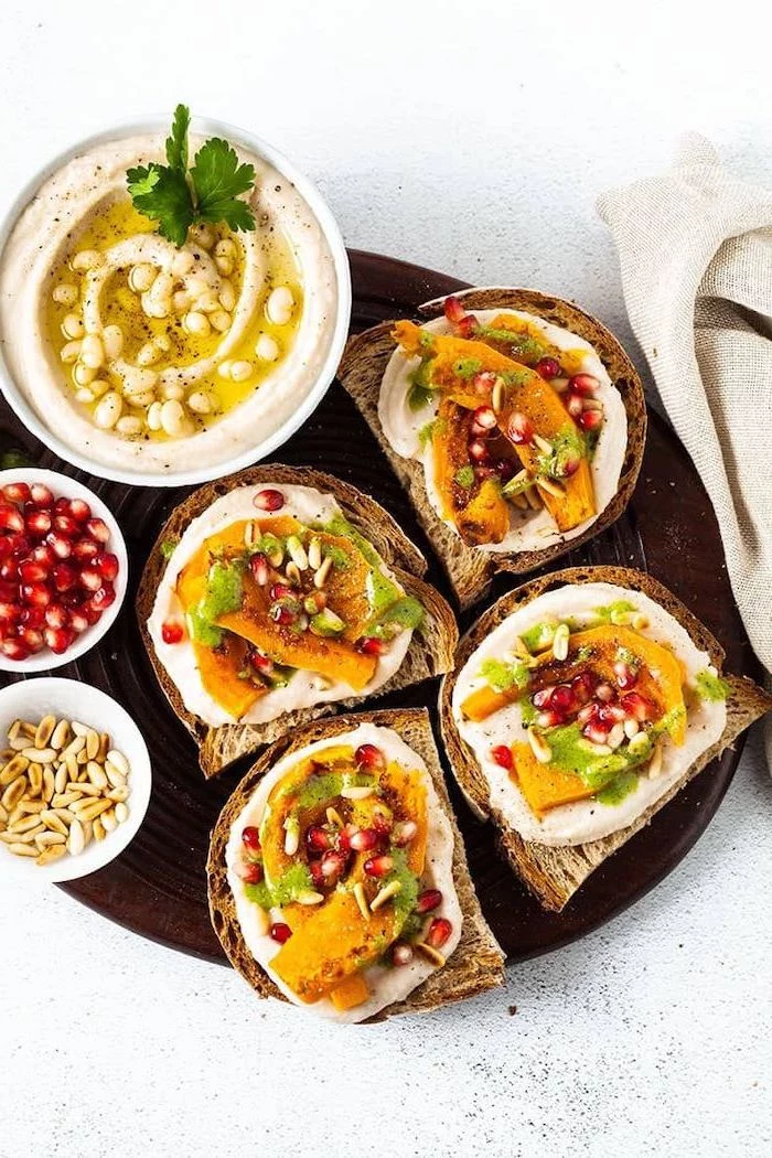 toast with sweet potato wedges hummus pomegranate seeds arranged on brown plate vegan appetizers white bowl full of hummus