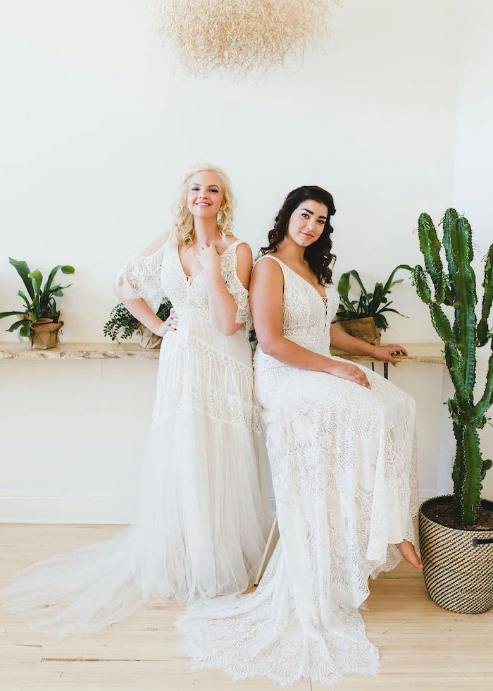 two women wearing all lace white unique wedding dresses sitting on tall chair green plants around them