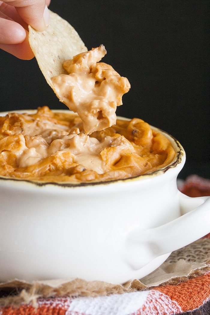 vegan party food baked hummus inside white bowl tortilla chips dipped into it held by male hand