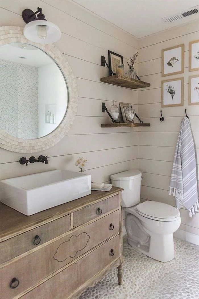 white shiplap on the walls cobble stone on the floor farmhouse bathroom decor wooden vanity round mirror above it two shelves above the toilet