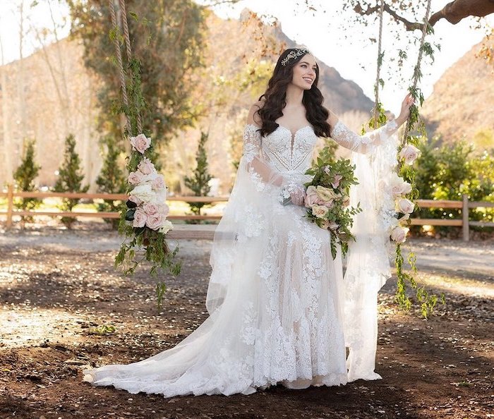 woman with long wavy black hair sitting on a wooden swing boho beach wedding dress wearing all white lacy dress with long sleeves