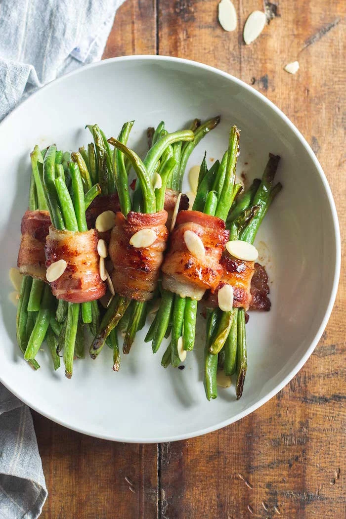 bacon wrapped green beans served on white plate with silvered almonds christmas food ideas placed on wooden surface