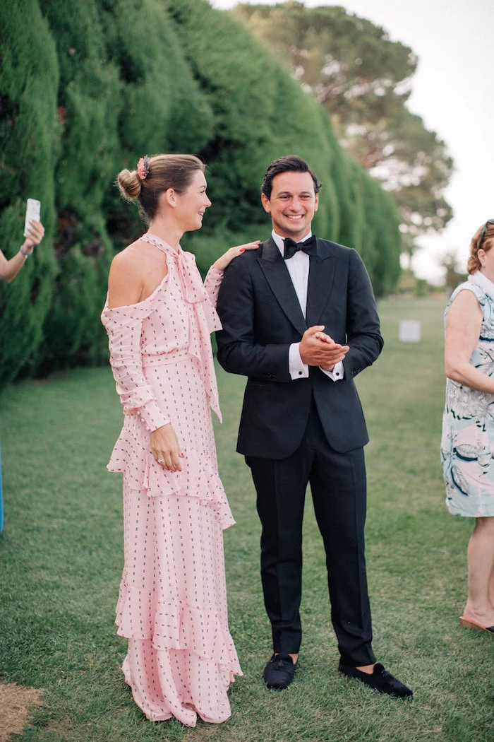 black suit worn by man standing next to woman wearing long pink dress with fringe black dots and long sleeves womens wedding guest dresses