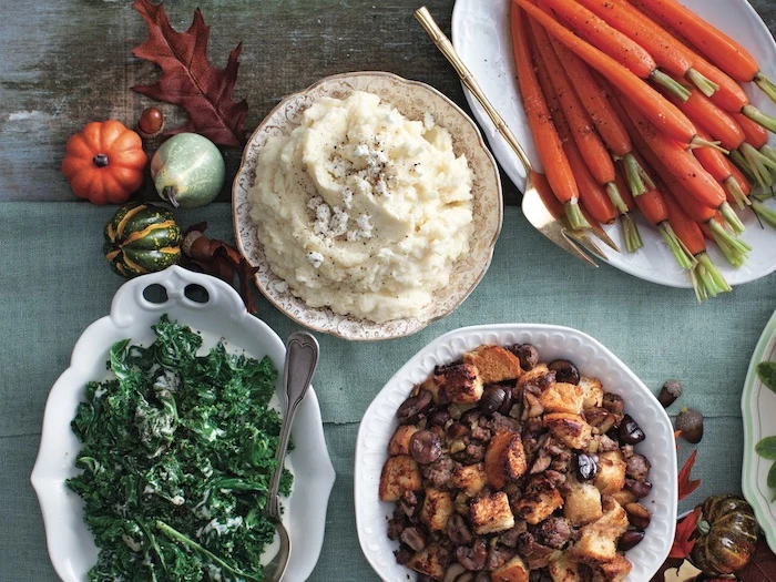 four thanksgiving side dishes on white plates on blue wooden surface mashed potatoes stuffing carrots kale salad