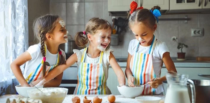 indoor activities for kids three girls wearing colorful aprons beating egss in white bowls flour on their faces