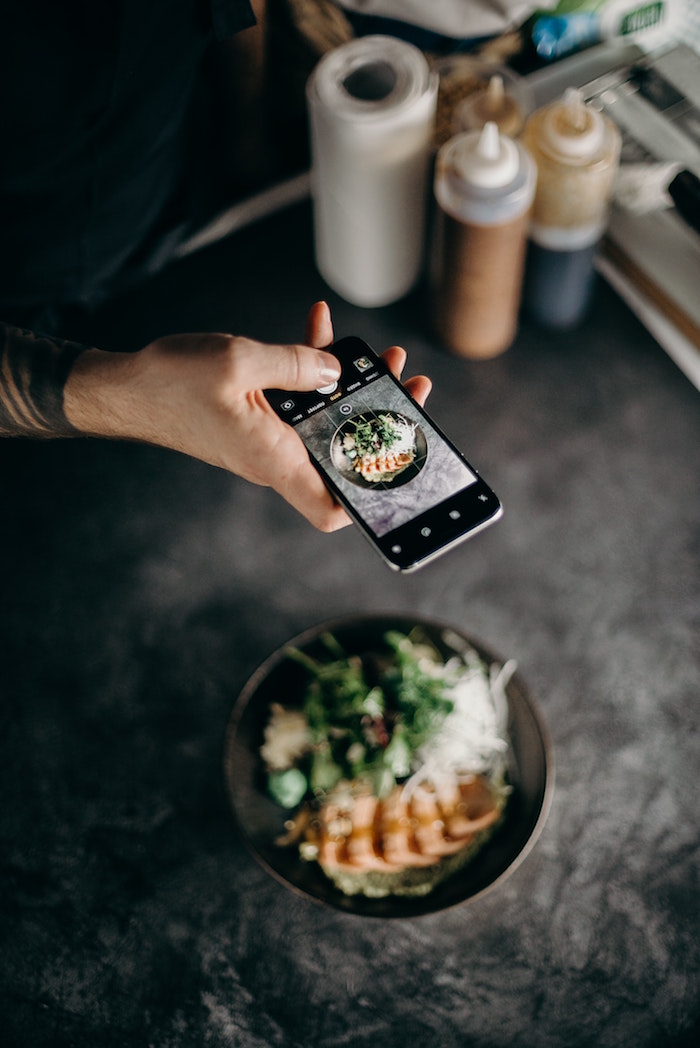 instagram followers hand holding phone taking picture of black plate filled with food placed on black surface