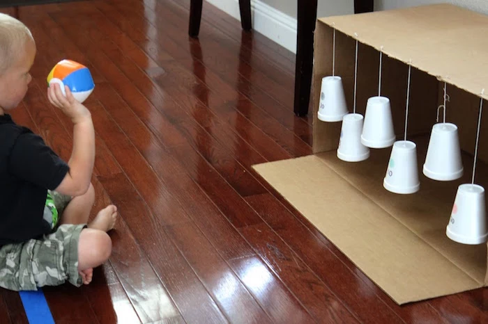 kid sitting on the floor throwing small ball at styrofoam cups hanging from carton box art and craft ideas for kids