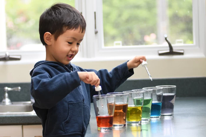 little boy wearing black hoodie standing next to kitchen counter indoor activities for kids six glasses filled with water in different colors
