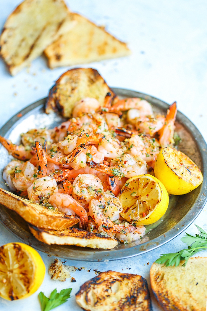 metal bowl filled with garlic butter shrimp garnished with herbs roasted lemon wedges toasted bread on the side