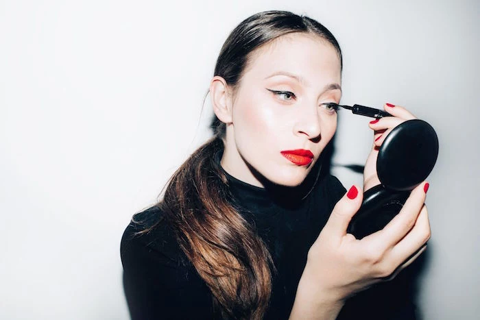 photo of woman putting eyeliner on her eyes holding a mirror how to do a cat eye wearing black blouse