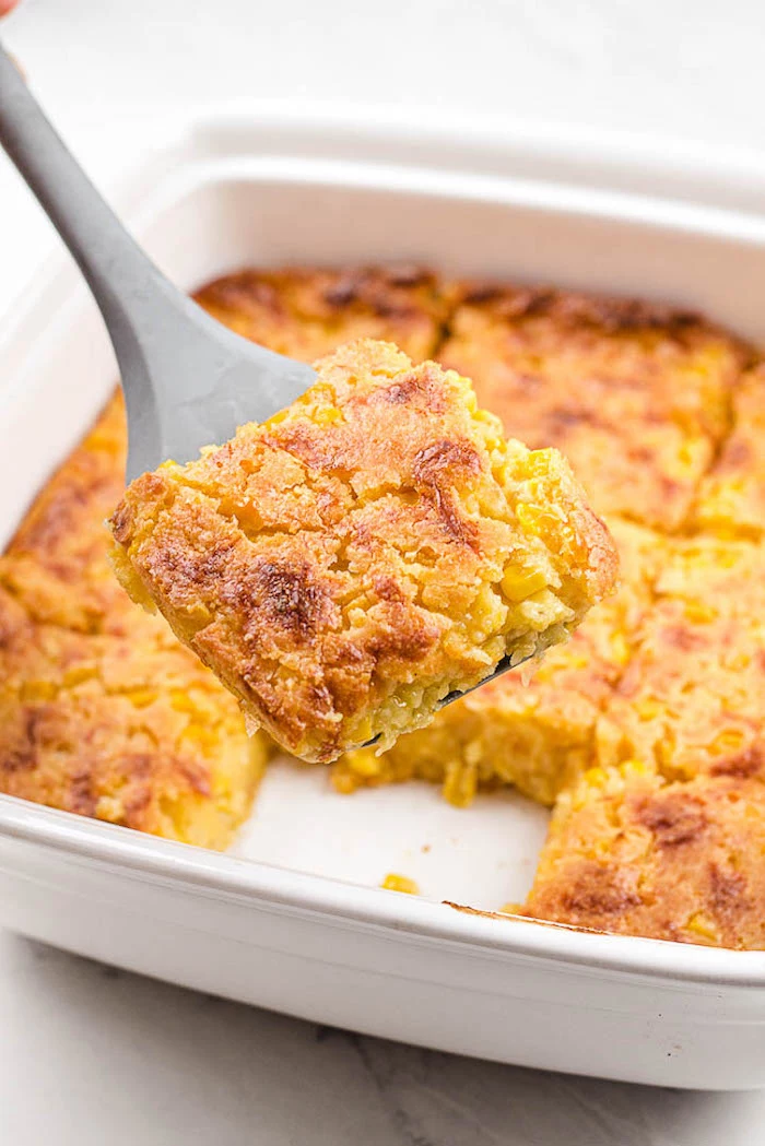 piece of corn casserole on spatula easy thanksgiving side dishes white casserole dish filled with corn in the background