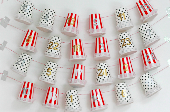 red white and black paper cups labeled with numbers hanging from red and white strings hanging on white wall homemade advent calendar