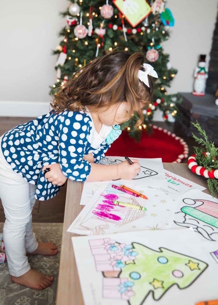 toddler coloring on wooden table wearing white pants blue blouse with white dots coloring sheets for kids christmas tree in the background
