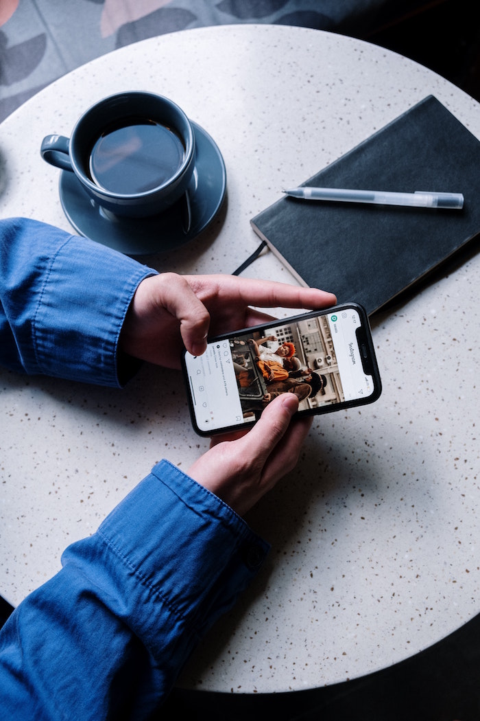 two hands holding phone scrolling instagram followers coffee mug notebook with pen on white round table