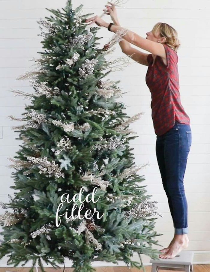 white background christmas tree theme ideas woman wearing red plaid shirt jeans standing on a stool adding filler to christmas tree christmas tree theme ideas