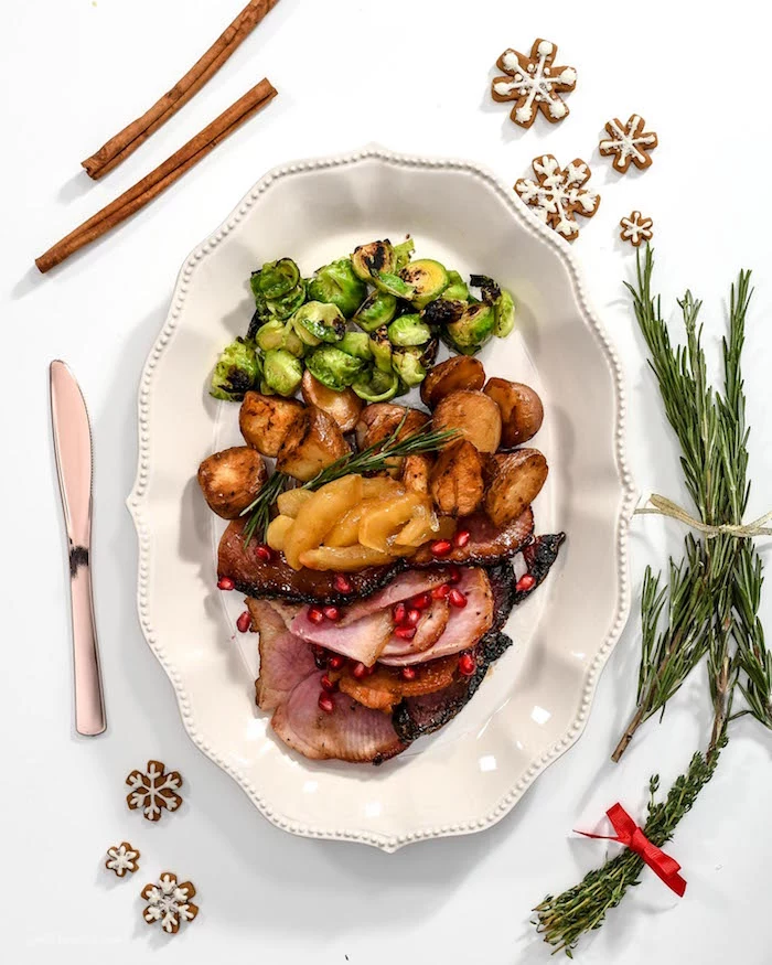white plate with slices of ham brussels sprouts potatoes placed on white surface christmas dinner 2020 knife rosemary branches cinnamon sticks snowflake cookies on the side
