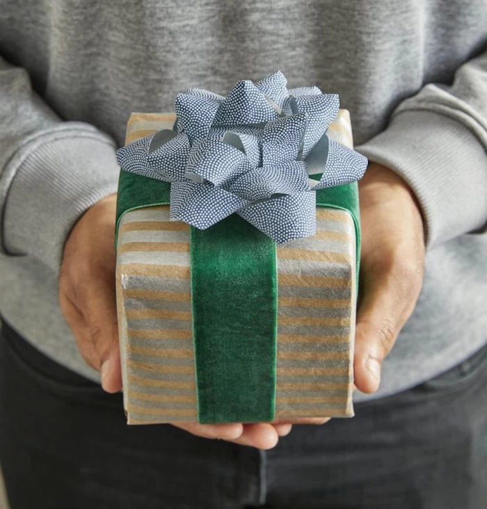 man wearing gray blouse black jeans holding a gift diagonal gift wrapping wrapped with gray and gold wrapping paper green velvet ribbon