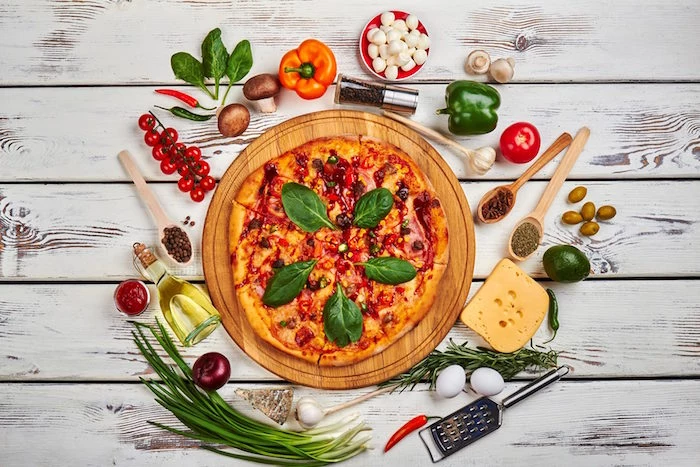pizza garnished with basil leaves placed on wooden cutting board homemade pizza dough different toppings around it