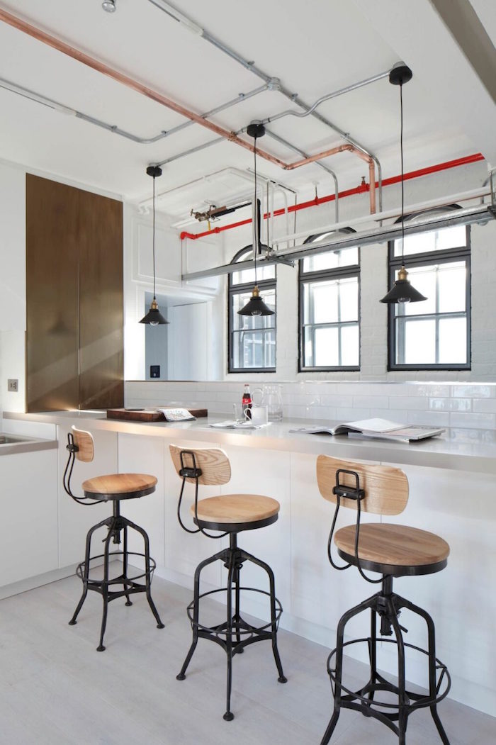 three wooden bar stools next to white countertop attached to wall covered with white subway tiles industrial pipes on the ceiling
