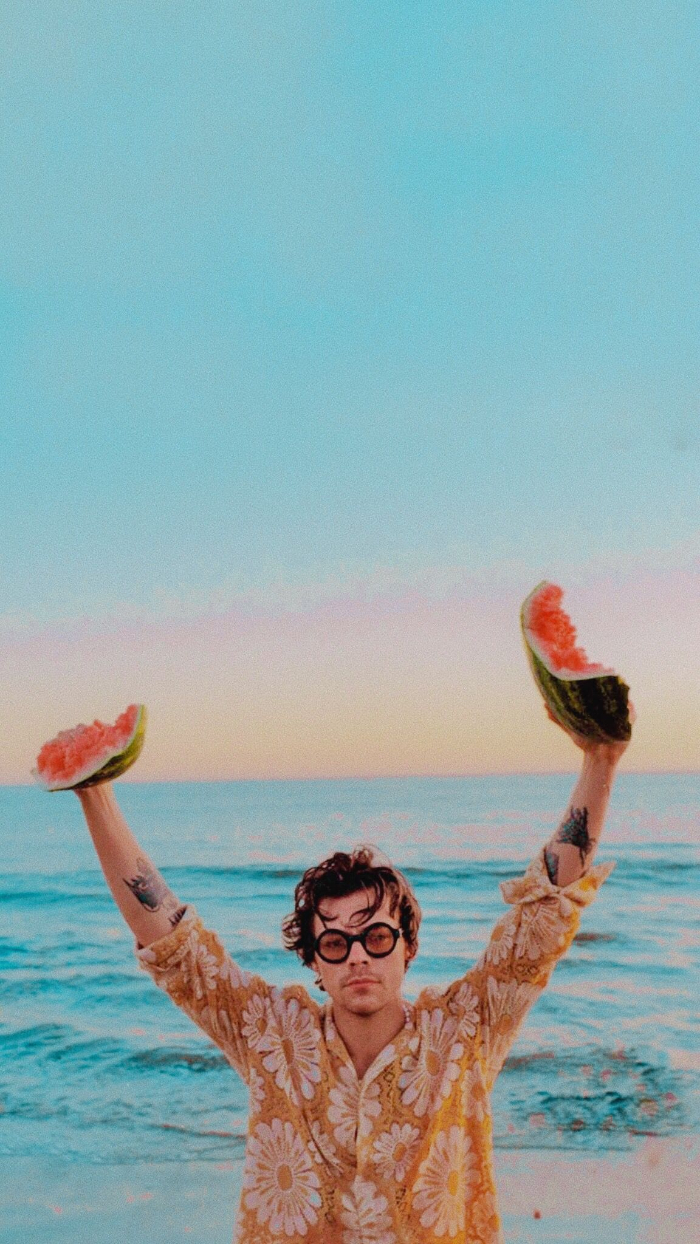 beach ocean waves in the background harry styles wallpaper wearing orange floral shirt holding pieces of watermelon