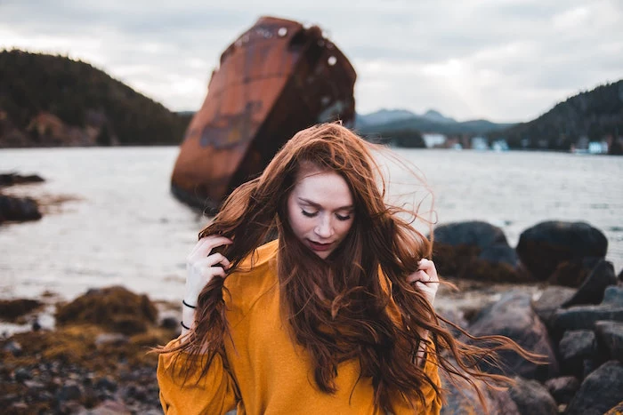 ginger haired woman wearing mustard yellow sweater standing next to lake keep your hair healthy long wavy hair