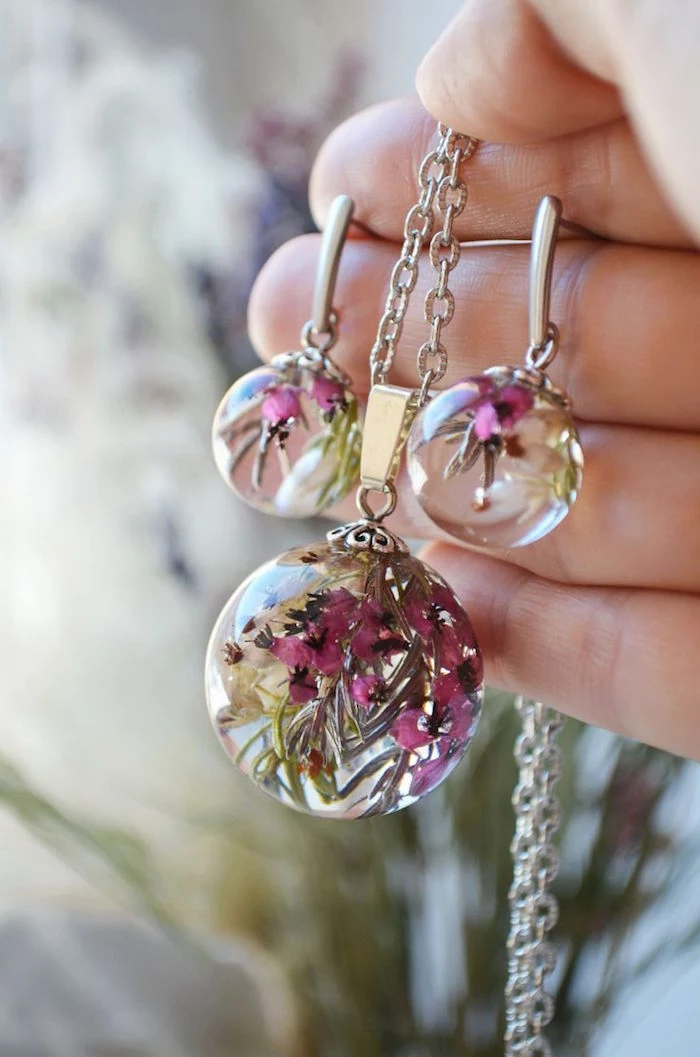 hand holding silver necklace and pair of earrings resin jewelry pink dried flowers inside blurred background