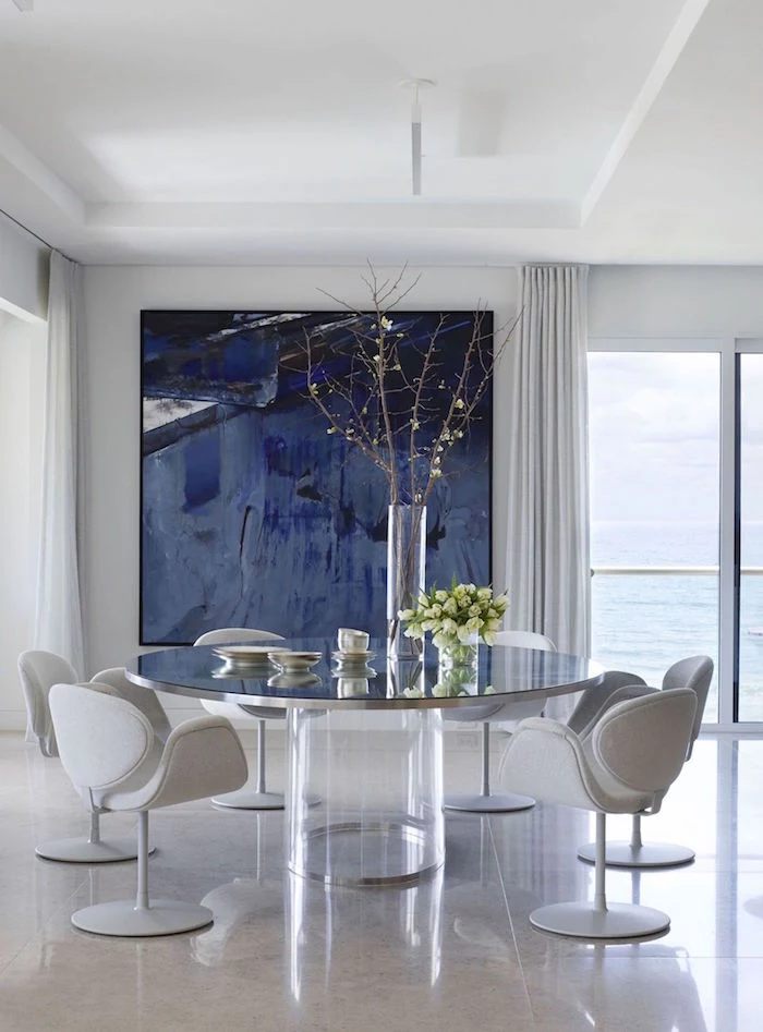 small beach house large round glass table with light gray chairs around it large abstract painting on the wall in shades of blue