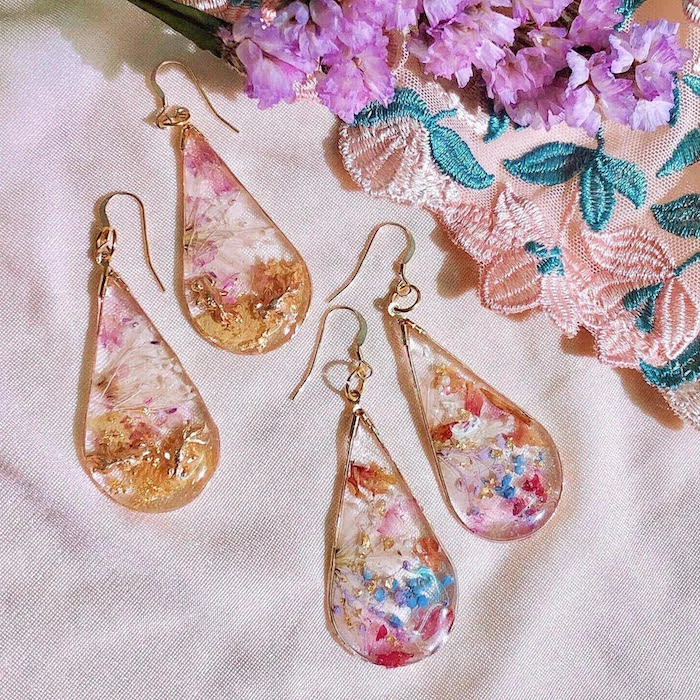 two pairs of earrings in raindrop form dried flowers on the inside in different colors resin jewelry placed on white silk cloth